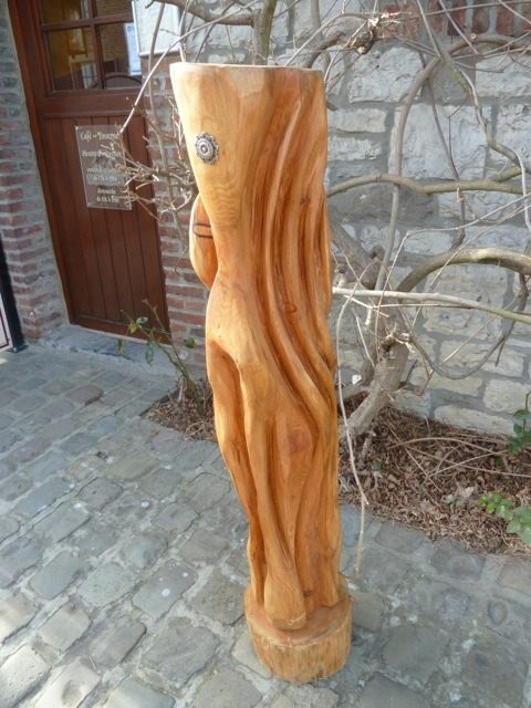 Totem orange aux plis ondulants, orné d'un œil en métal, devant une porte en bois.